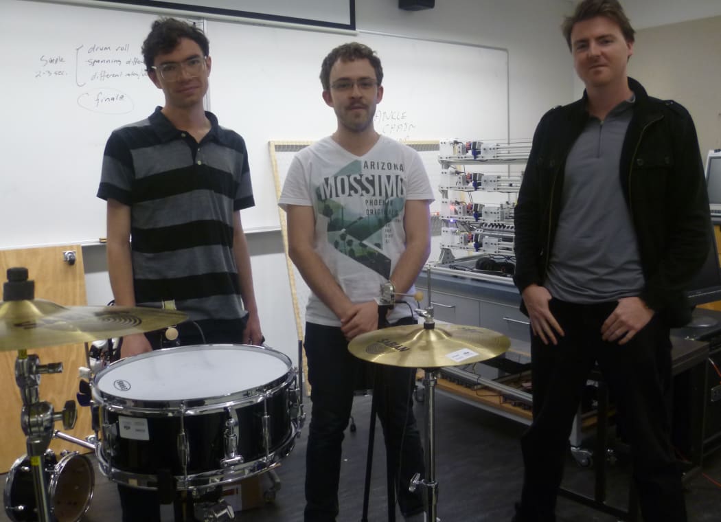 Jim Murphy (left), James McVay (centre) and Jason Long (right) standing behind the robotic drum kit. The MechBass robotic bass guitar is at the back, between James and Jason.