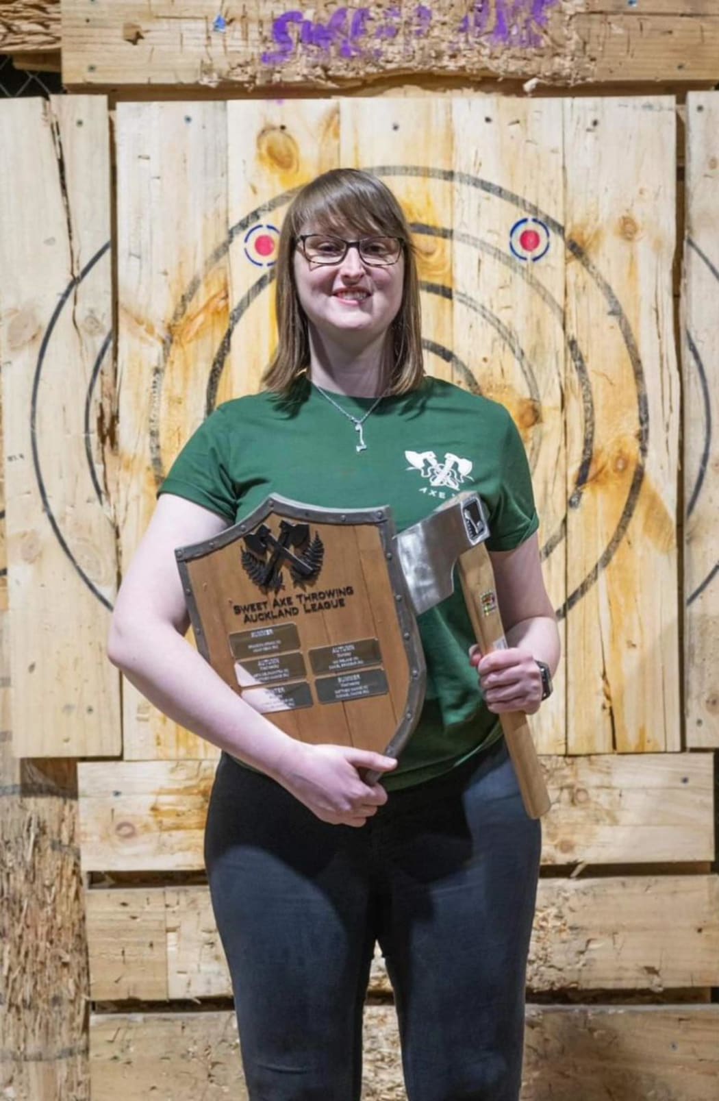 Britt stands in front of a target wall. She holds a trophy in one hand and an axe in the other.