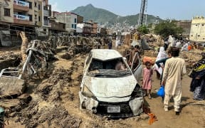People gather near a damaged vehicle and scattered debris after the road washed out following a flash flood in Mingora, the main city of Swat Valley, in monsoon-hit northern Pakistan's mountainous Khyber Pakhtunkhwa province on August 16, 2025. The death toll from heavy monsoon rains that have triggered flash floods across northern Pakistan has risen to at least 321 people in the last 48 hours, disaster agencies said on August 16. (Photo by Mehboob UL HAQ / AFP)