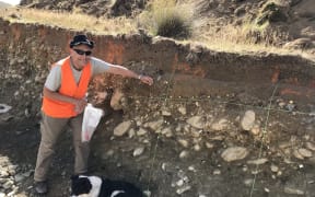 Mark, in high vis vest, sunglasses and hat places a plastic tag in the trench wall. His dog Sage is at his feet.