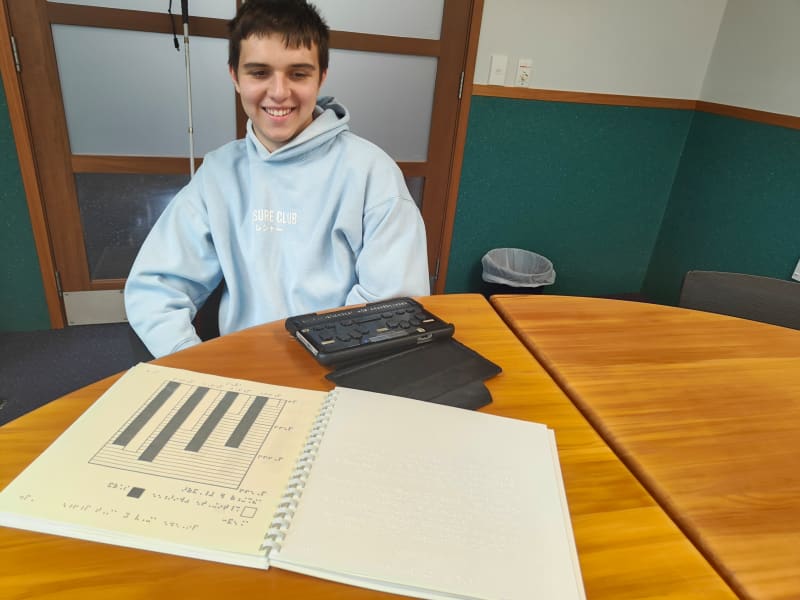 Toby Ireland smiles as he sits in front of a desk with a booklet showing a graph and his BrailleSense device.