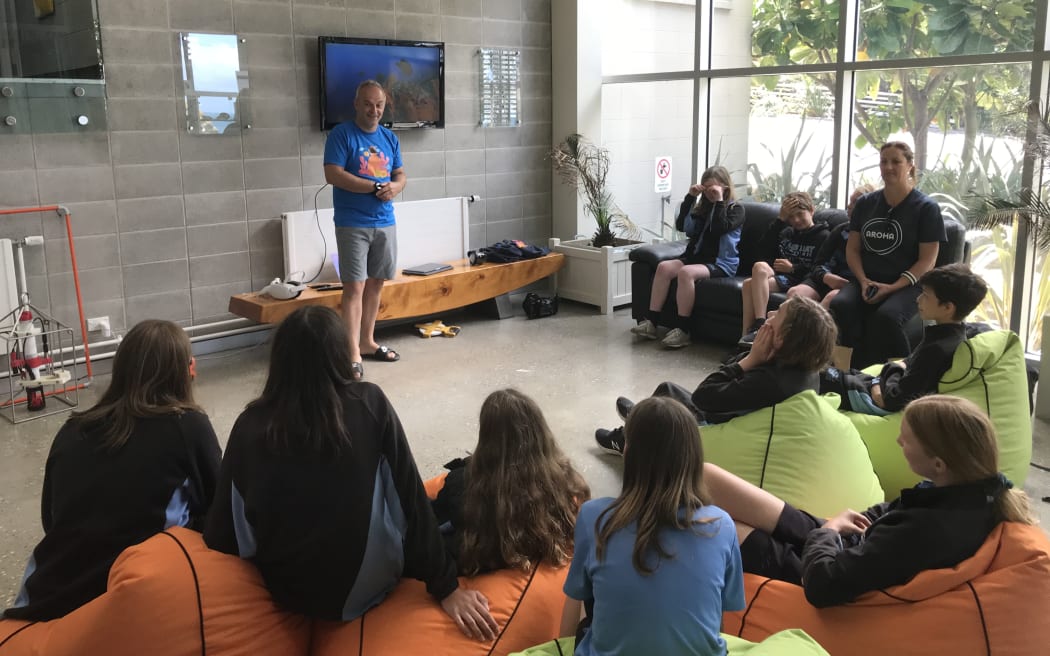 A group of students sit on coloured bean bags in a semi-circle listening to Professor James Bell give a presentation. They are in a big foyer area. Behind James is a screen.