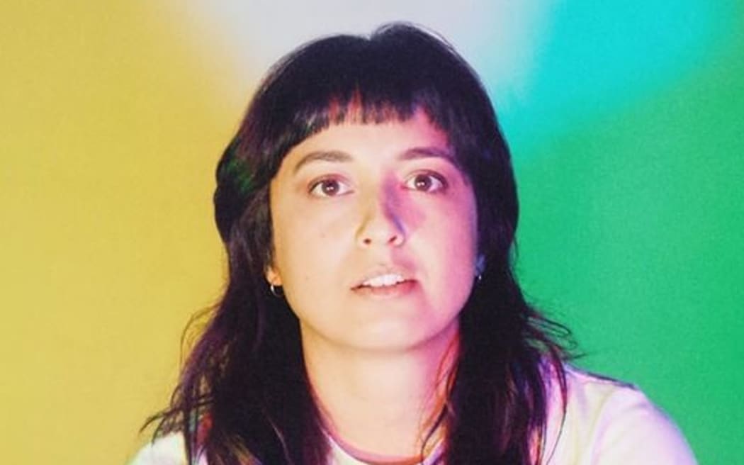 Liz Stokes - a young woman with long brown hair - gazes at the camera with a colourful background behind her.
