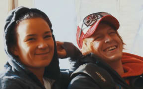 Two smiling young people - a woman wears a hoodie and man wears a trucker cap.