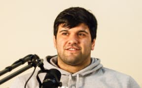 A man wearing a grey hoodie half smiles, in front of him is a microphone.
