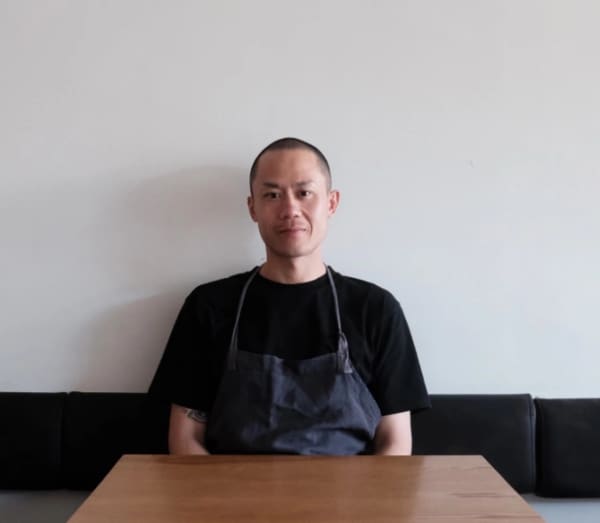 Chef John Yip sits at a table wearing a black t-shirt and a dark coloured apron.