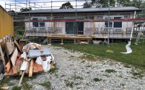 A small house is under construction on a plot of grassy land. There is scaffolding around the structure and the exterior walls are not yet finished. There is a shipping container to the right with building materials strewn around it. It is a cloudy day.