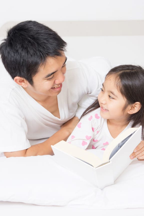 A photo of a little girl reading a story book with her father on the bed