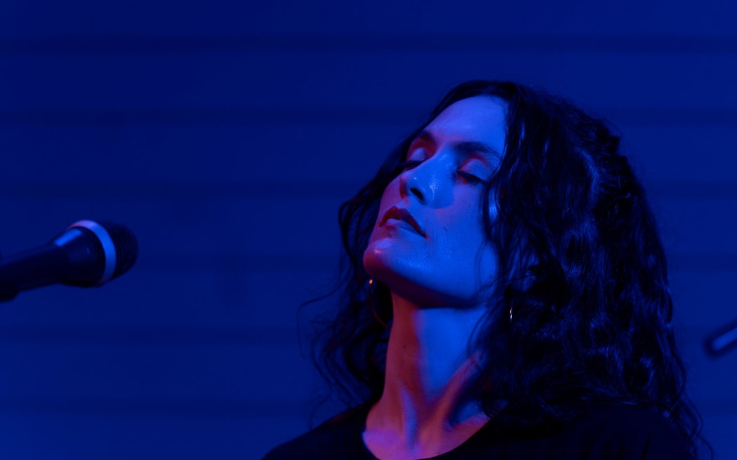 Babe Martin stands onstage under blue light.
