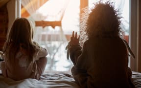 Back view of kids in warm costumes sitting together on bed in warm sunlight