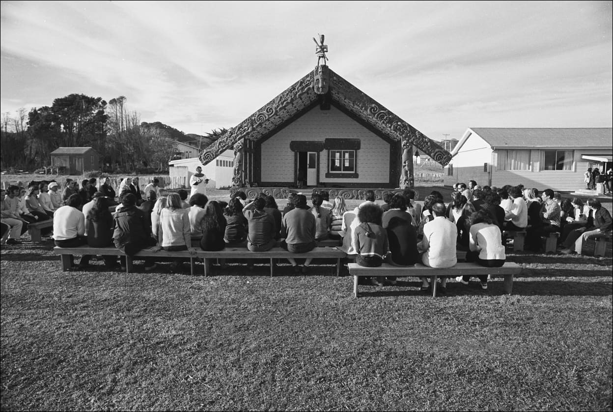 A black and white photo of a large group seated on benches outdoors, facing a marae.