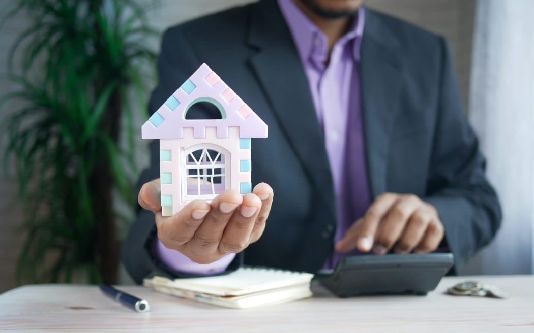 A man in a suit holding a model of a house calculates a sum.