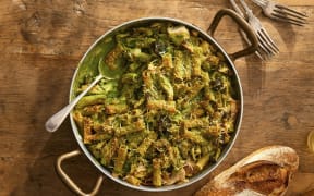 A round dish of chicken pasta bake on a wooden board, with forks and a baguette in shot.