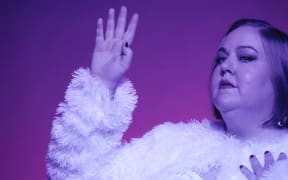 Aubrey Gordon - A fat woman with blonde hair stands unsmiling holding her right hand aloft. She is wearing a striped dress and a fluffy jacket whilst bathed in purple light and set against a pink and purple background.