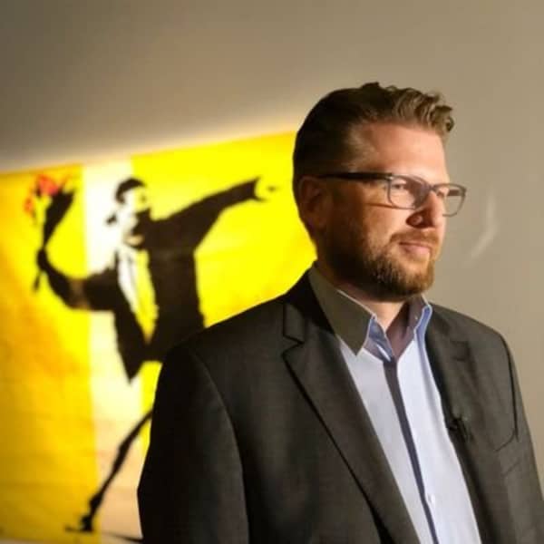 A bearded man in a suit stands in front of a brightly colured artwork.