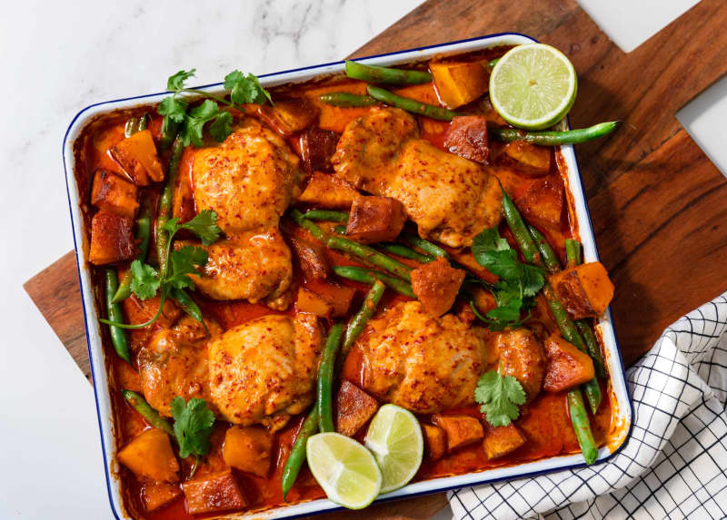 A rectangular oven tray containing red curry chicken and pumpkin tray bake, with green beans, coriander leaves and lime wedges.