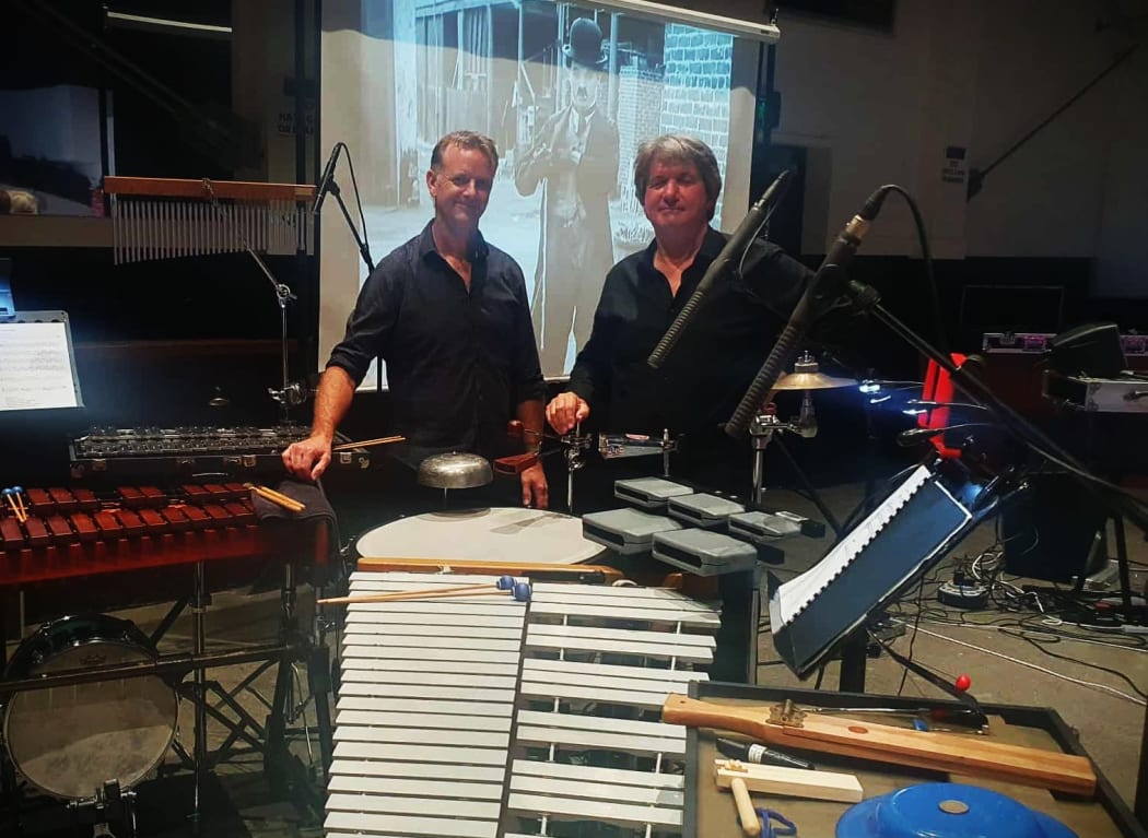 David and Jeremy stand behind a mass of instruments. In the background is a projector showing a silent film.