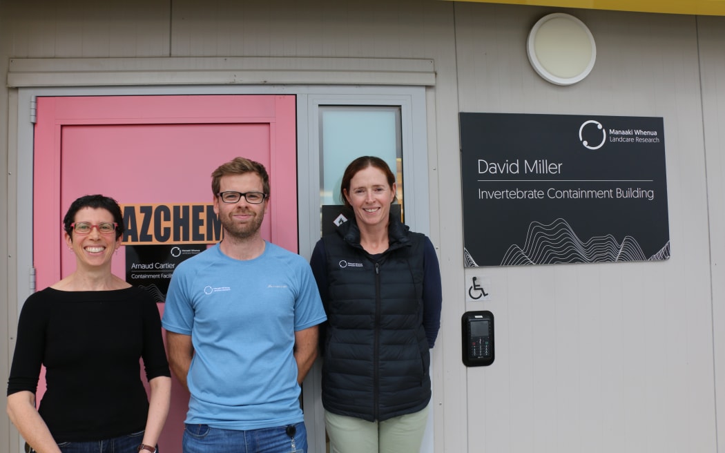Three people stand in front of the door to the Invertebrate Containment Facility, smiling at the camera.