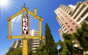 For rent sign inside ruler in shape of house, with high rise building in background.