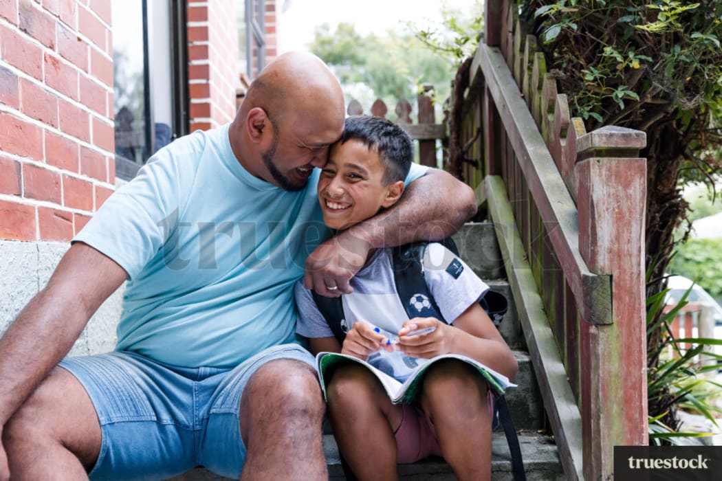 A stock photo of a father and son sitting on the steps outside a house, smiling. The father embraces the son.