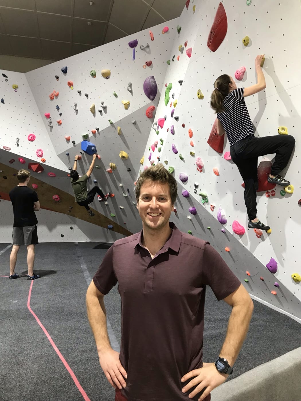 Patrick Boudreau at Resistance Climbing Gym. Patrick stands in front of a bouldering wall with two climbers up on the walls and one waiting.