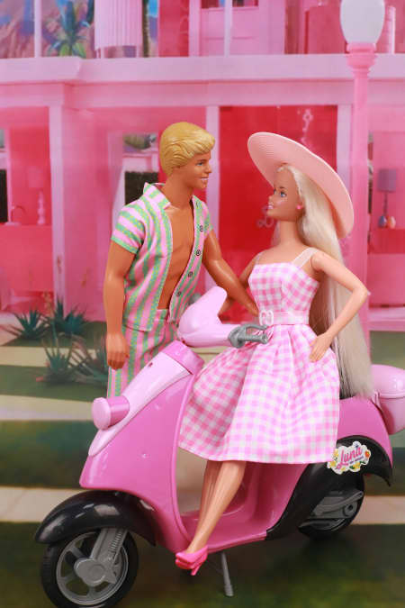 A Ken doll faces a Barbie doll, who wears a pink dress and a pink hat and sits on a pink motorbike.