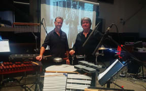 David and Jeremy stand behind a mass of instruments. In the background is a projector showing a silent film.