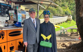 Jacqui and her husband stand outside a food truck in their vintage clothes. Jacqui wears a navy cape, blue and yellow tartan skirt, yellow gloves, a green bag, and a matching tartan bonnet.
