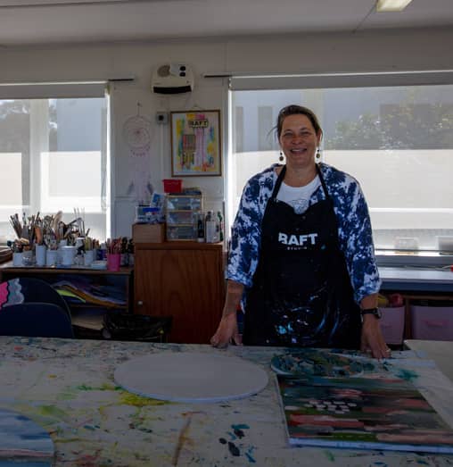 Raft Studios founder Jen van der Woerd stands smiling in her workshop behind a table with her painting.
