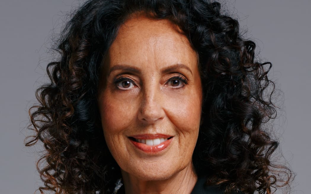 Headshot image of Moana Maniapoto smiling at the camera in front of a grey background.