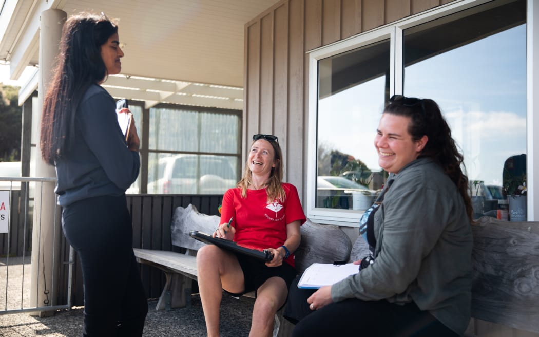 Sanaa Nair, Lydia Green and Catherine Meyer laugh together sitting outside a small airport terminal.