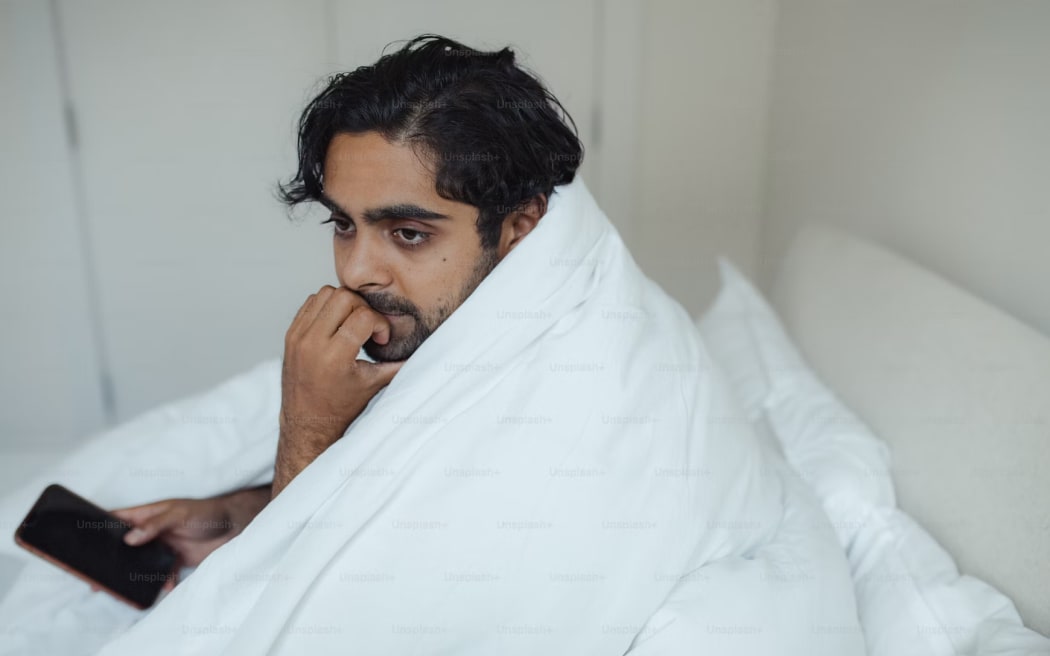 Un hombre en la cama con un teléfono celular y una mirada preocupada.