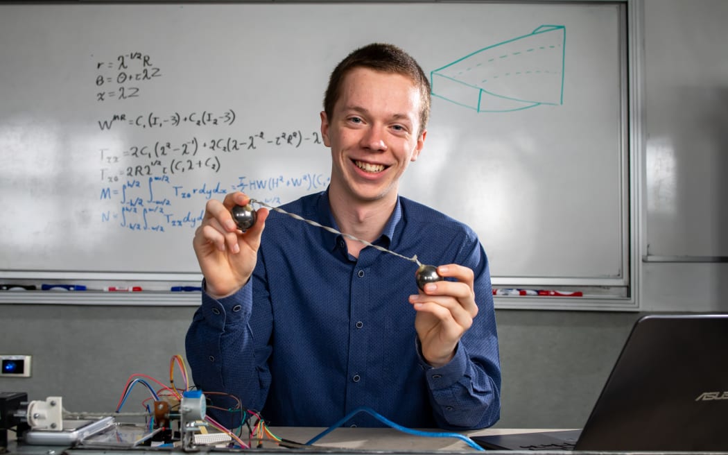 A young man sits at a desk in front of a whiteboard covered with mathematical equations. He is holding a twisted rubber band.