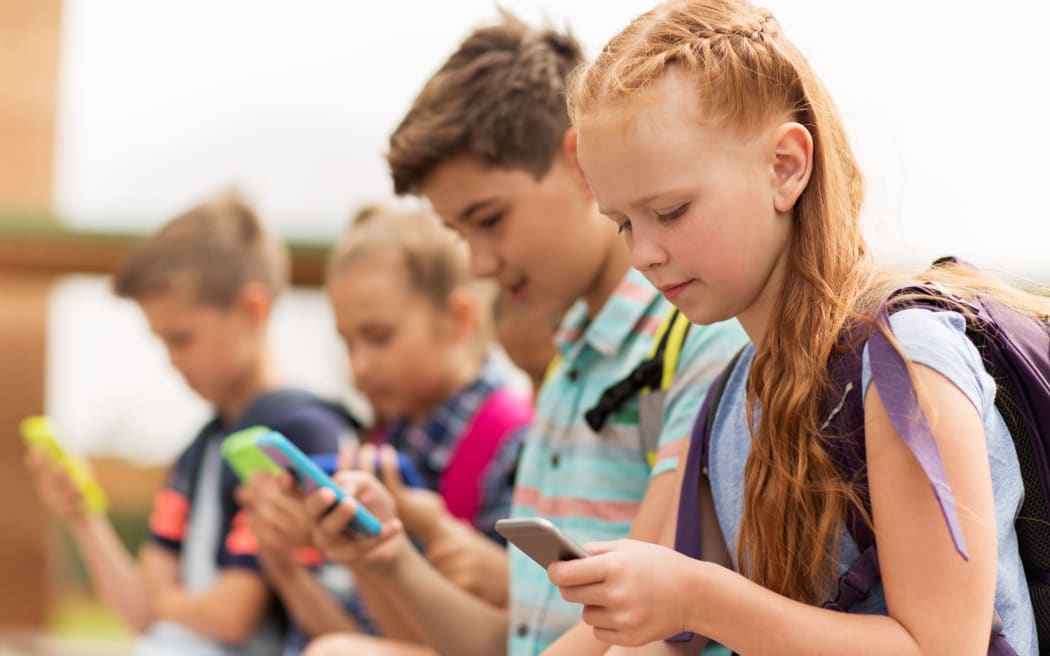 primary education, friendship, childhood, technology and people concept - group of happy elementary school students with smartphones and backpacks sitting on bench outdoors