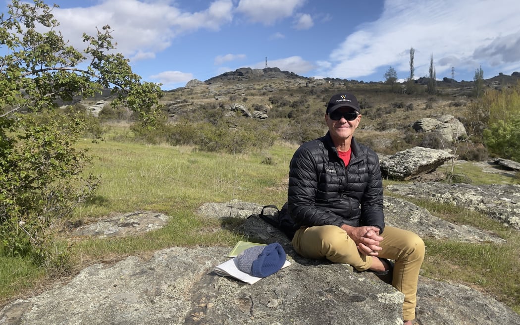 A man in a cap, sunglasses, black pufferjacket and brown pants sitting on a rock in a dry scrubby hillscape dotted with rocks. The sky is blue with puffy clouds.