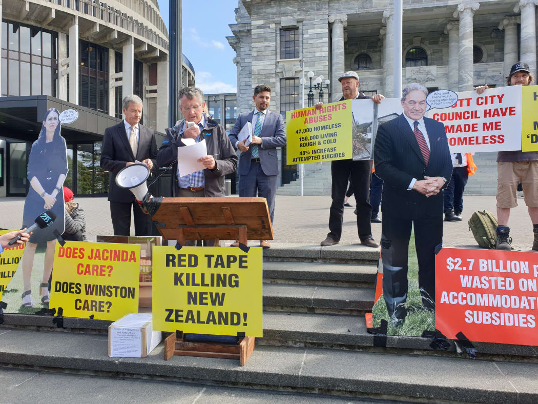 Colin Wightman speaking in a microphone at the grounds of Parliament in protest for legislation to protect tiny houses.