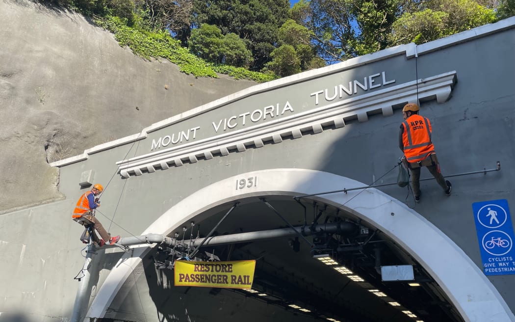 Two protesters from the Restore Passenger Rail group have abseiled down the Hataitai side of the Mt Victoria Tunnel in Wellington and hung a sign saying 'Restore passenger rail'.