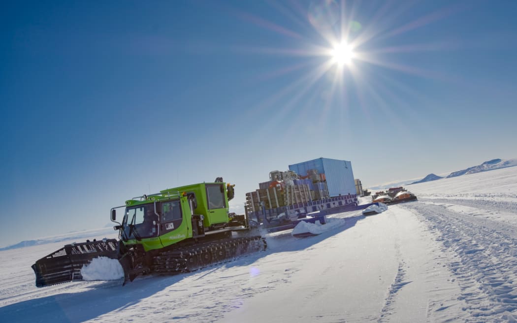 A lime-green heavy vehicle on caterpillar tracks makes its way across a snowy plain in Antartica, towing a large sled laden with freight containers.