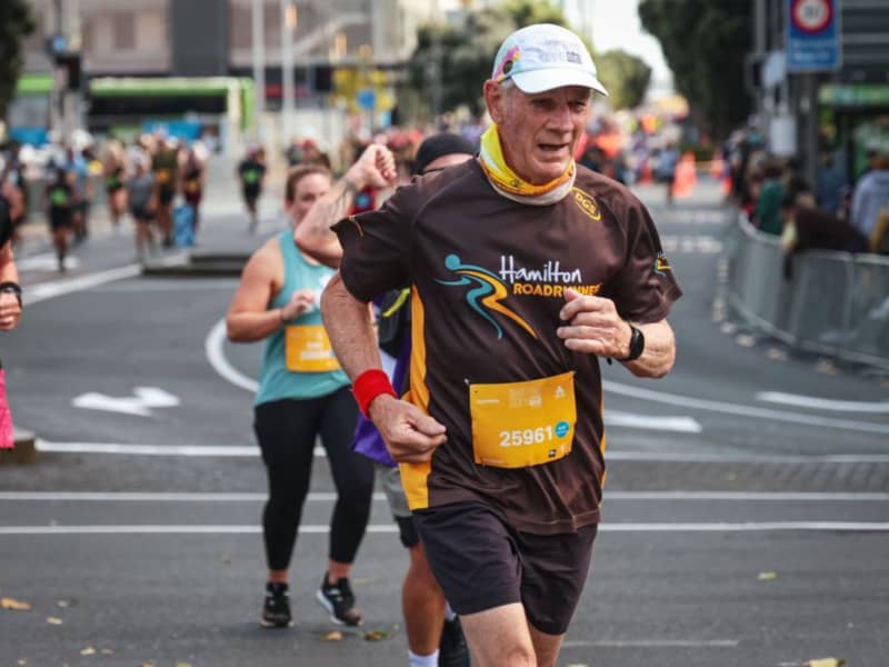 Rod Gill - a wearing brown shorts and t-shirt and white cap - runs ahead of a group of others along a road.