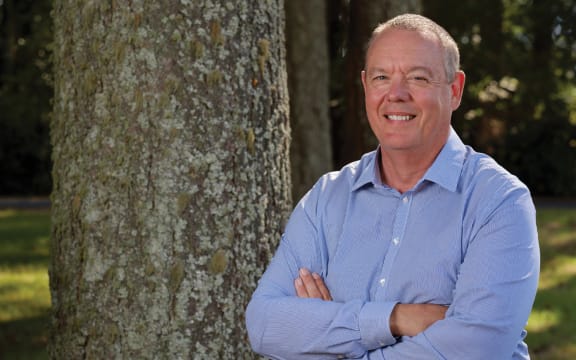 A headshot of Julian standing with his arms folded in front of a tree. He is smiling and wearing a blue button-up shirt.