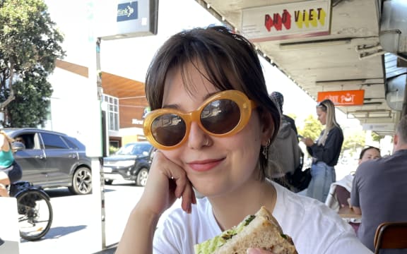 Bryer is sitting at a cafe table on a busy Wellington street. She smiles at the camera, wearing funky yellow sunglasses and holding half of a sandwich.