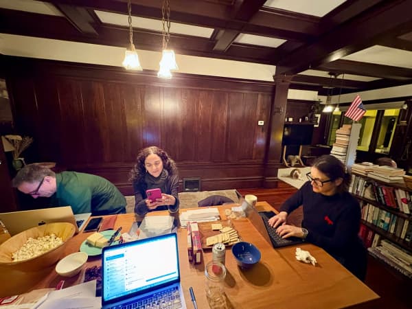 A group of adults gathered in a lounge on a dining table, where there's a bowl of popcorn and people have their laptops out.