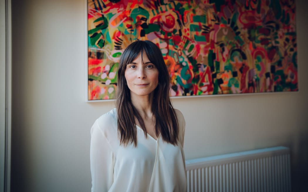 This is a shot of Josie from the waist up. She is standing in front of a radiator. Above that a large colourful painting hangs. She is wearing a white top and has slightly below shoulder length brown hair.