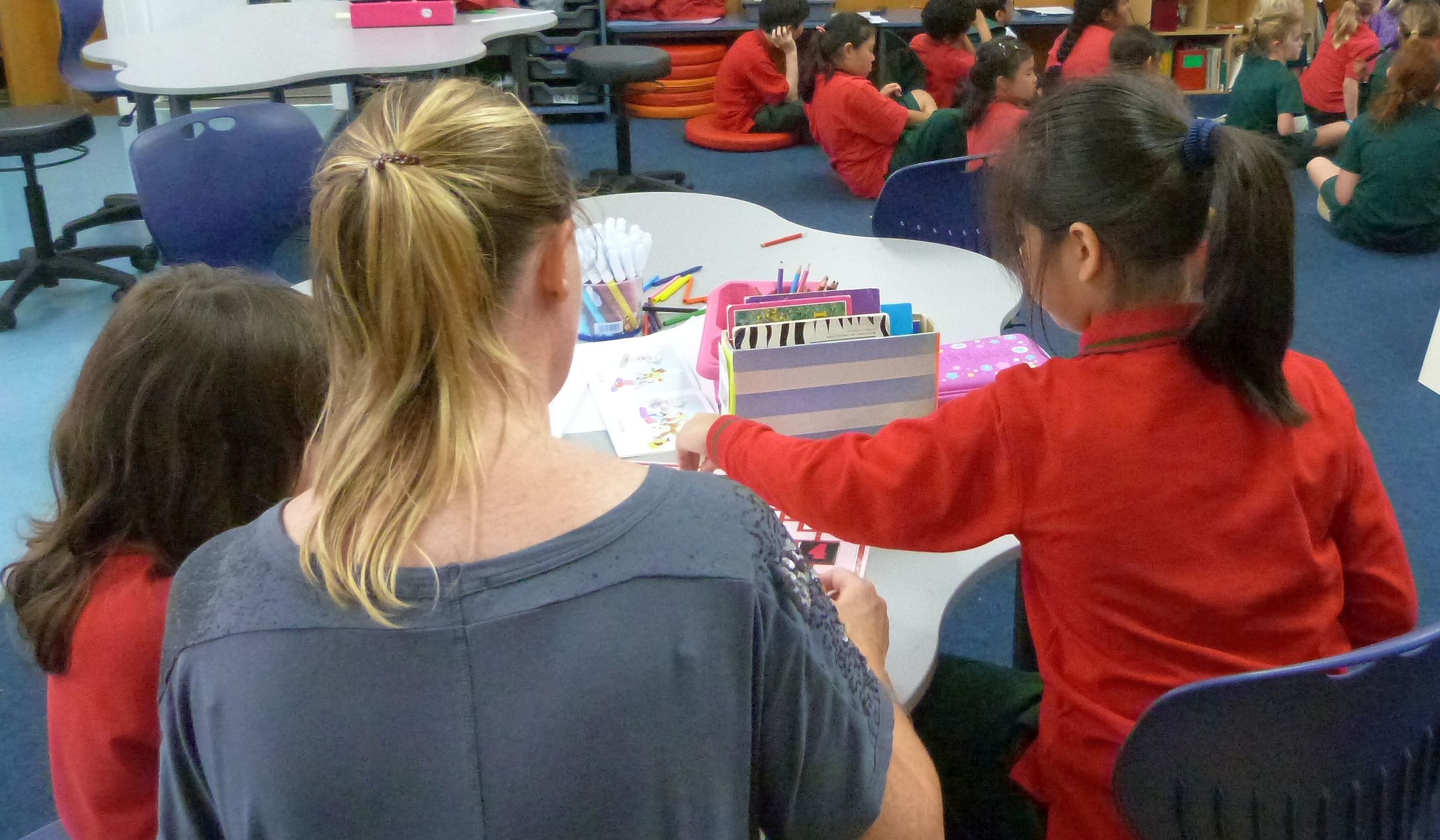 A view from behind of teacher aide and two little girls while rest of class taught  further into classroom.