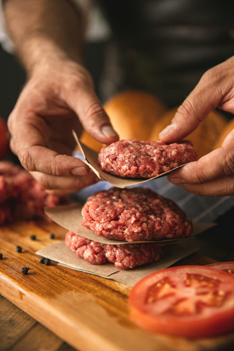 A pair of hands holds a minced meat burger pattie on a piece of baking paper, stacking it above two others on a board. Sliced tomatoes are in the foreground.