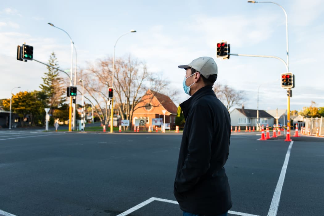 Man wearing a facemask waiting at the intersection for the green pedestrian light. Roadwork traffic cones along the street.