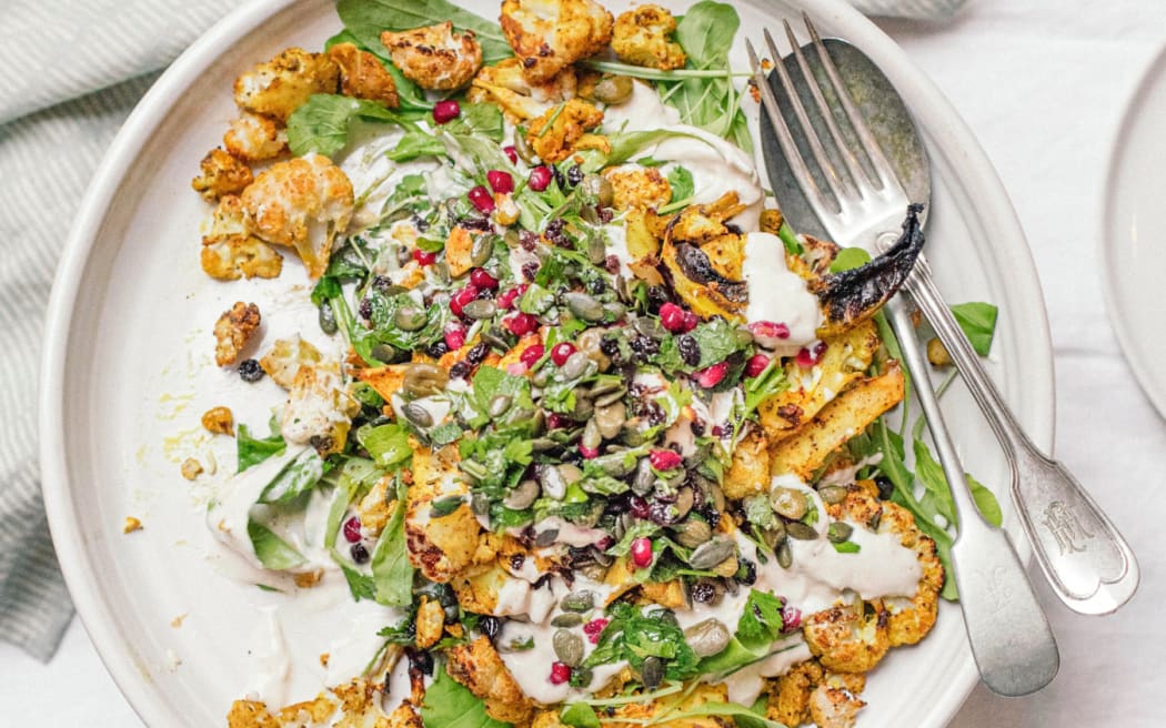 A white plate atop a white table, with a fork and spoon sitting nested on the side. The salad is piled on the plate: yellow roasted cauliflower, creamy sauce and bright zingy greens.