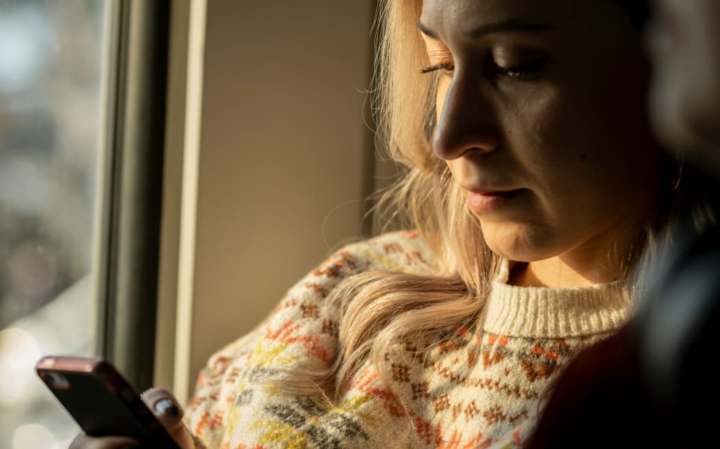 A blonde woman in soft light looks down at her phone.