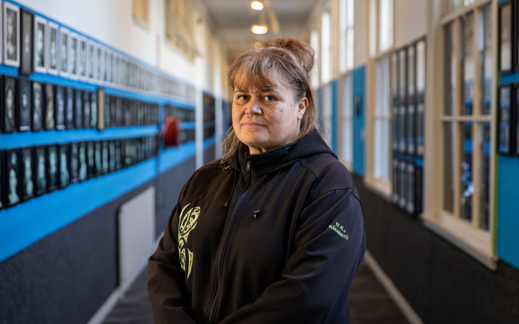 Te Pā ō Rākaihautū Principal Whaea Terina Tahau stands in the hallway holding her hands in front of her. She is wearing a black jacket and is looking directly at the camera. Behind her on the walls are framed pictures of students dressed in feather korowai or cloaks, with moko on their faces.