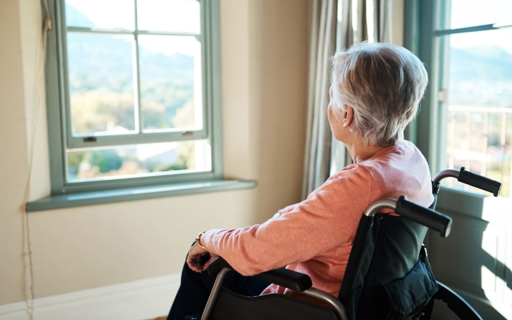 A senior woman in a wheelchair looking thoughtfully out of a window at home